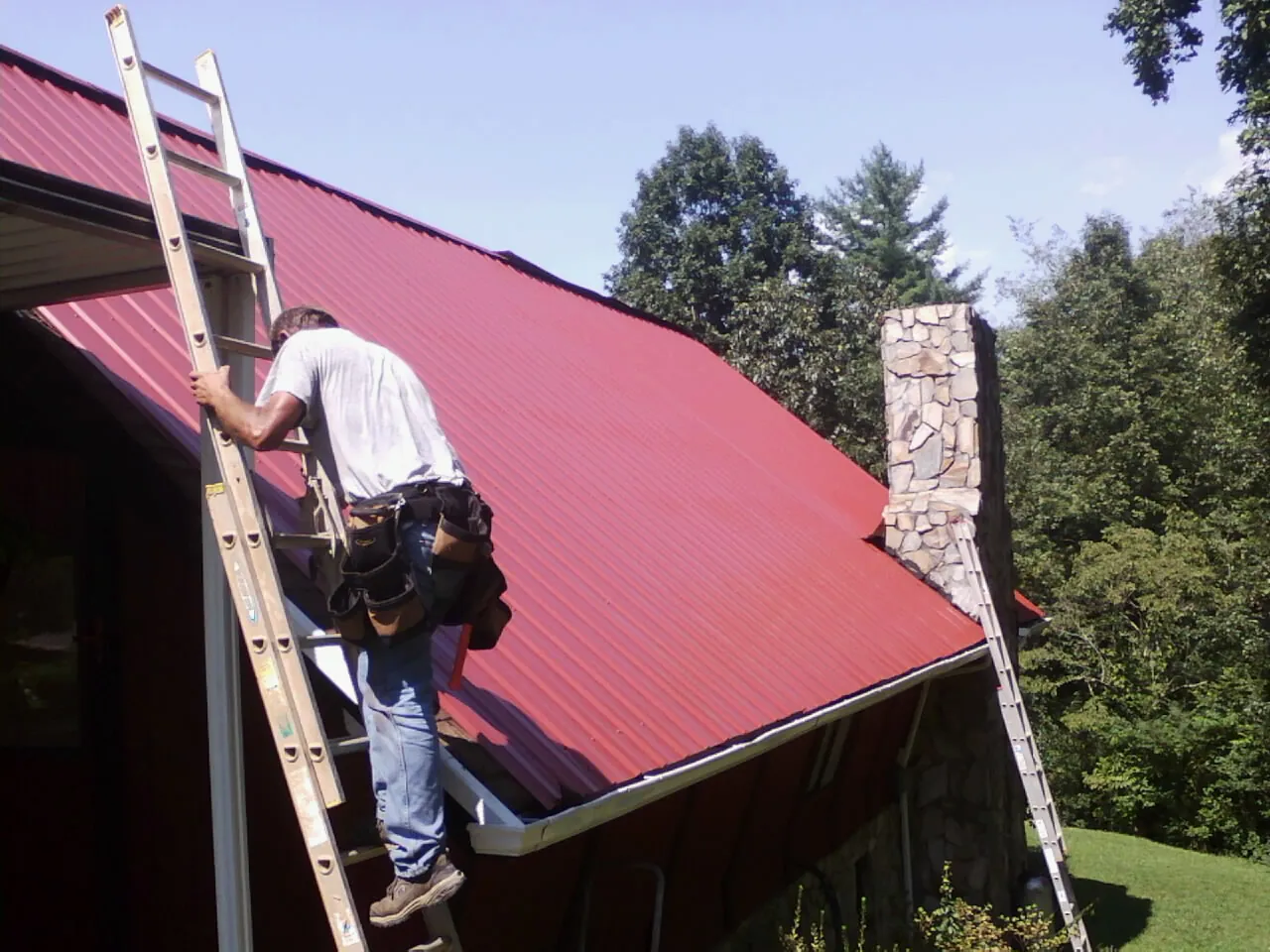 Roofer on a ladder beside a red metal roof and stone chimney — Soffit & Fascia Repair by Roofing Brothers in Royersford
